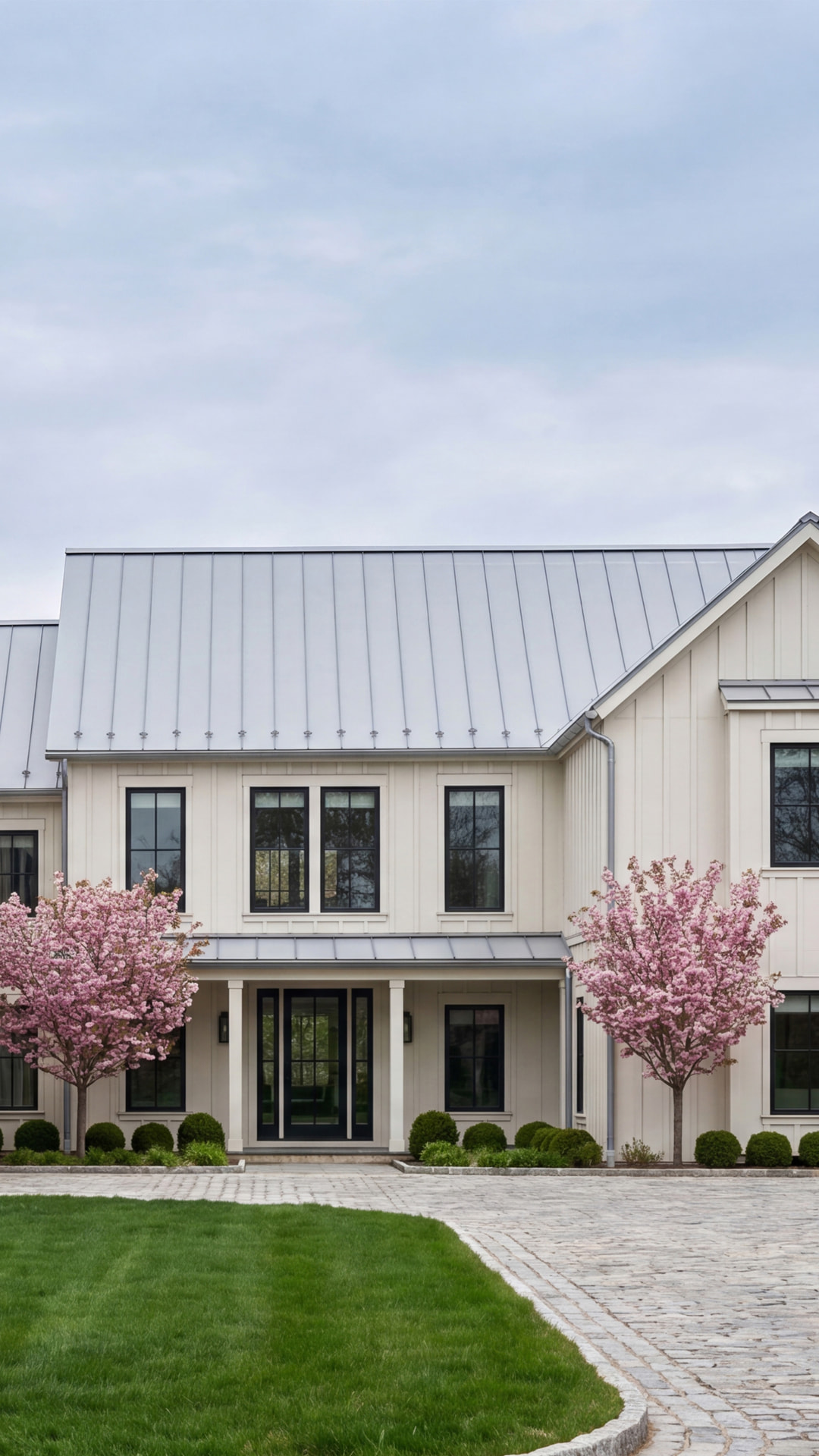 Completed board-and-batten residence with standing-seam metal roof, flowering cherry trees, and a cobblestone courtyard at handover.